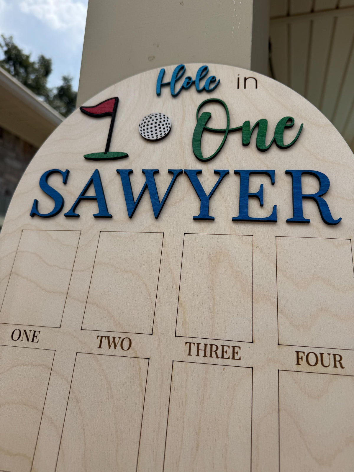 May include: Wooden golf-themed sign with the words "Hole in One" and the name "Sawyer" in blue and green lettering. Includes a golf ball and flag graphic. Features spaces labeled "One," "Two," "Three," and "Four."