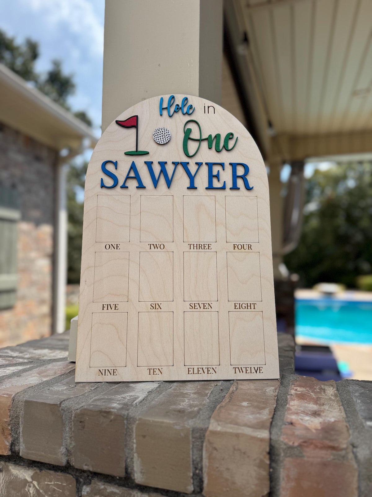 May include: Wooden golf score board with the text "Hole in One SAWYER". The top features a golf flag, golf ball, and the number 1. Below are 12 rectangular slots labeled "ONE" through "TWELVE".