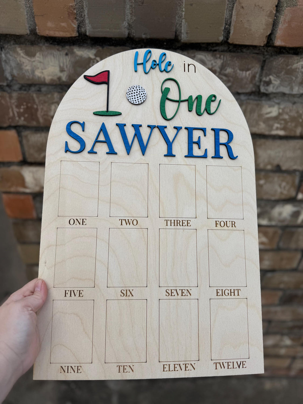 May include: Wooden golf-themed sign with the words "Hole in One" and "Sawyer" in blue and green. The sign features a golf ball and flag graphic. It has twelve rectangular spaces labeled "One" through "Twelve".