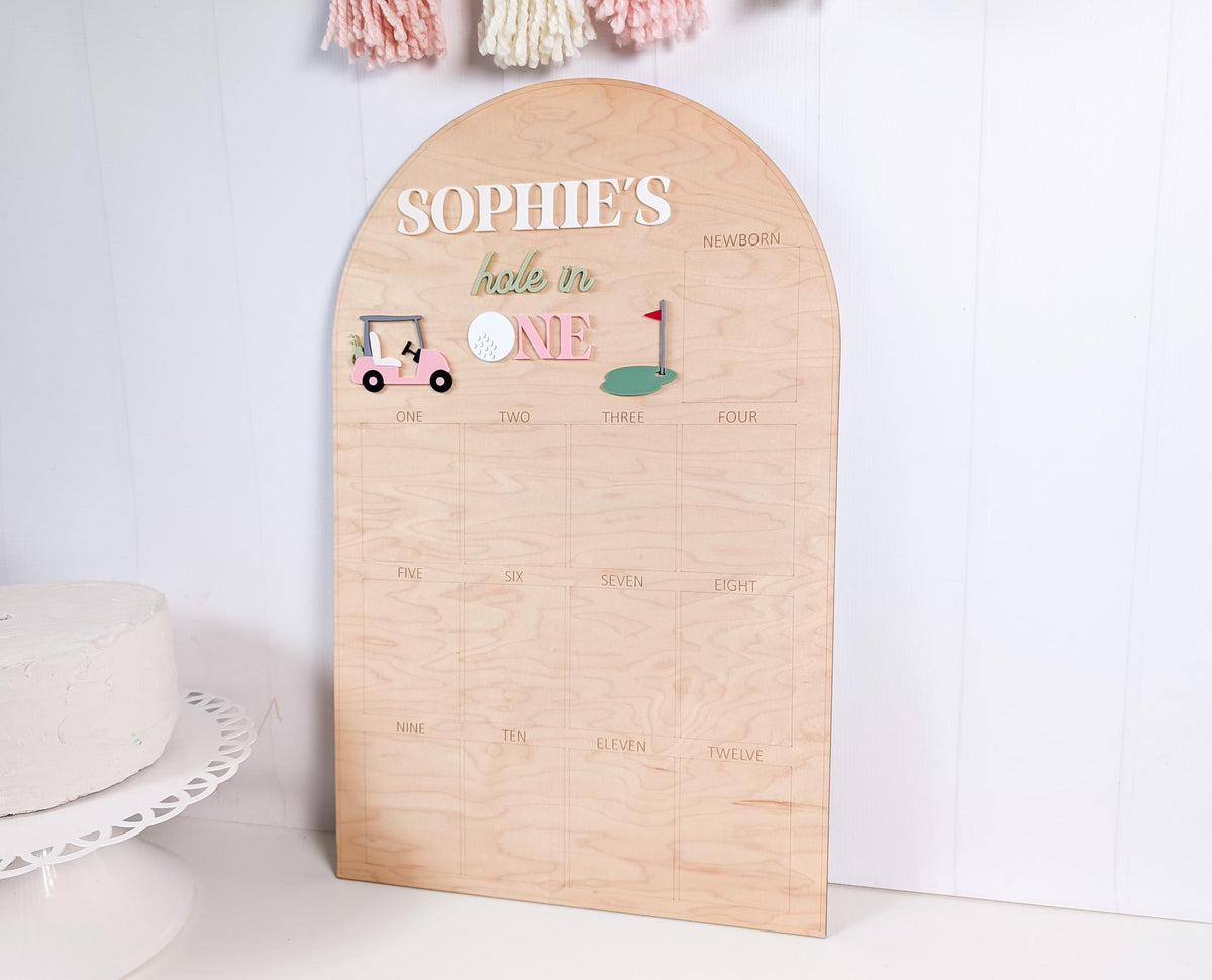 May include: A wooden golf-themed milestone board with an arched top. The board features the text "SOPHIE'S hole in ONE" in white and pink lettering, along with golf-related graphics. Numbers one through twelve are listed below the graphics.