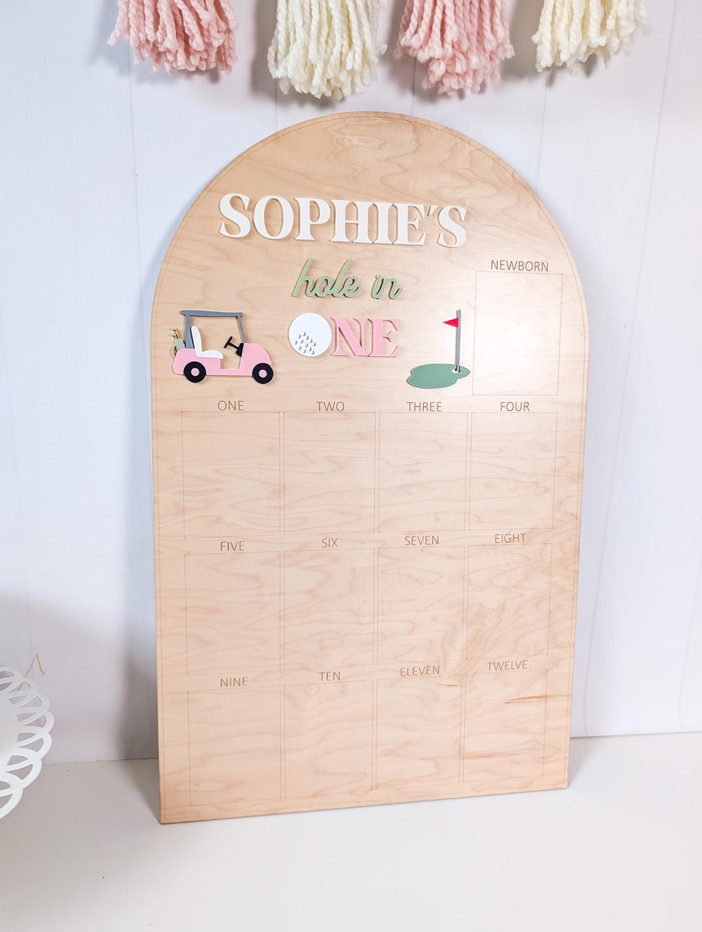 May include: A wooden sign with the text "SOPHIE'S hole in ONE" and a golf theme. The sign features a pink golf cart, a golf ball, and a green putting green. The sign has a grid with numbers one through twelve.