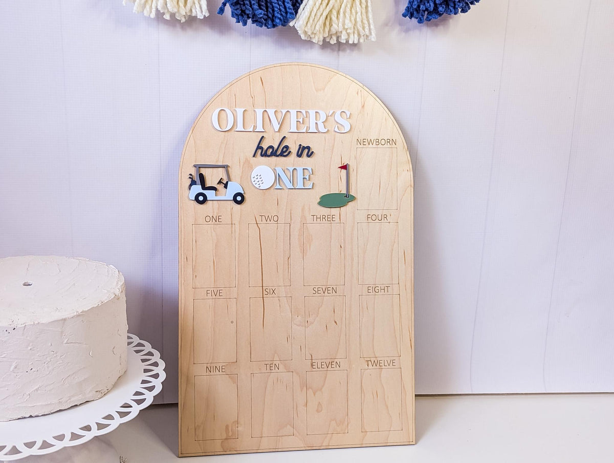May include: A wooden golf-themed milestone board with the text "OLIVER'S hole in ONE". It features a golf cart, golf ball, and a green with a flag. The board has spaces for tracking milestones from newborn to twelve.