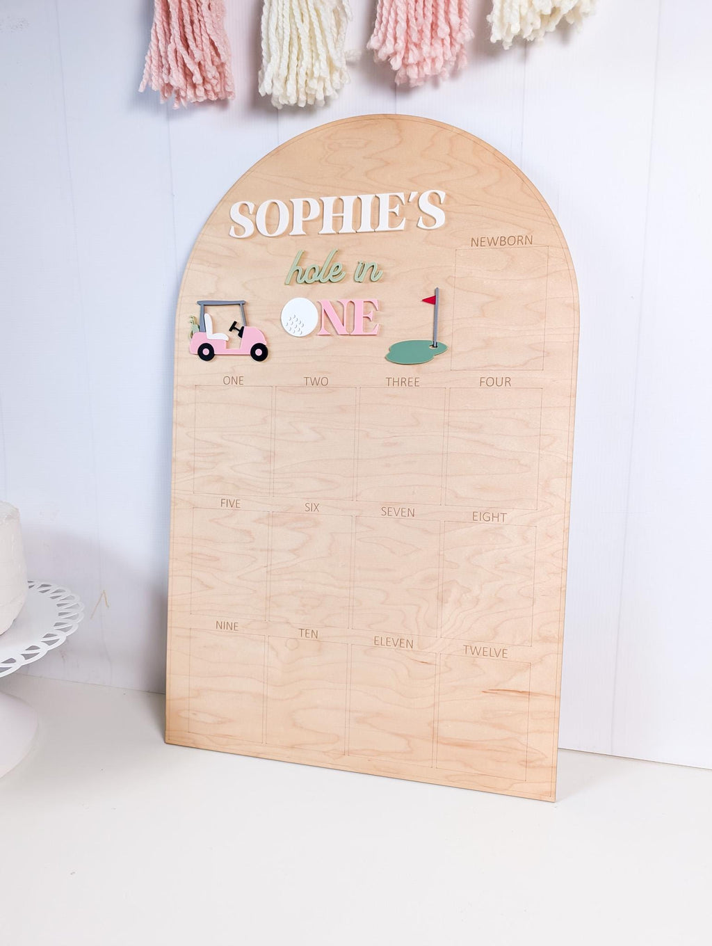 May include: Wooden milestone board with an arched top. The board features the text "SOPHIE'S hole in ONE" in white and green lettering, along with golf-themed illustrations. The board is divided into sections labeled "Newborn" through "Twelve".