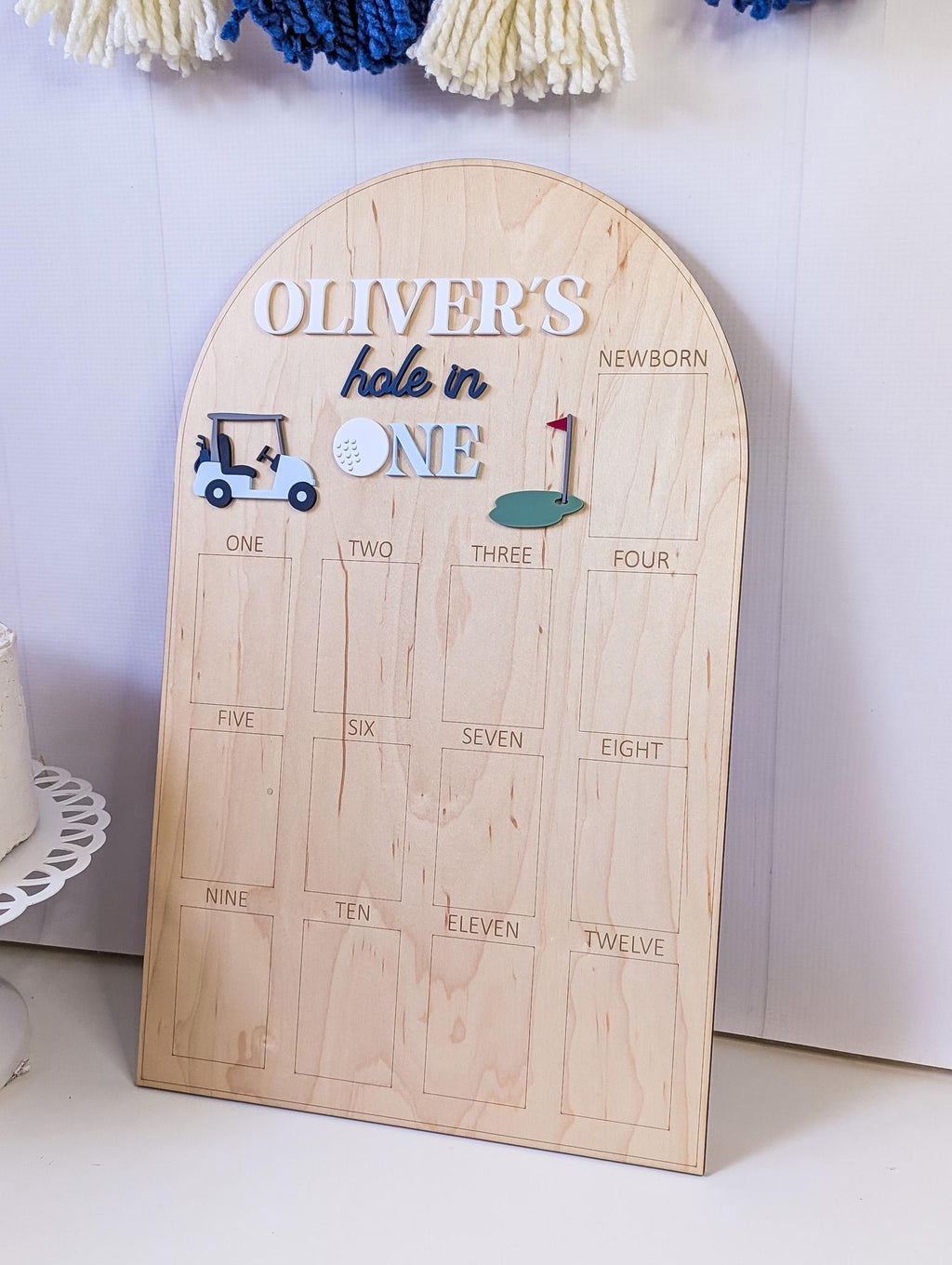 May include: Wooden milestone board with the text "OLIVER'S hole in ONE". The board features a golf cart, golf ball, and a green with a flag. It has spaces for tracking milestones from newborn to twelve months.