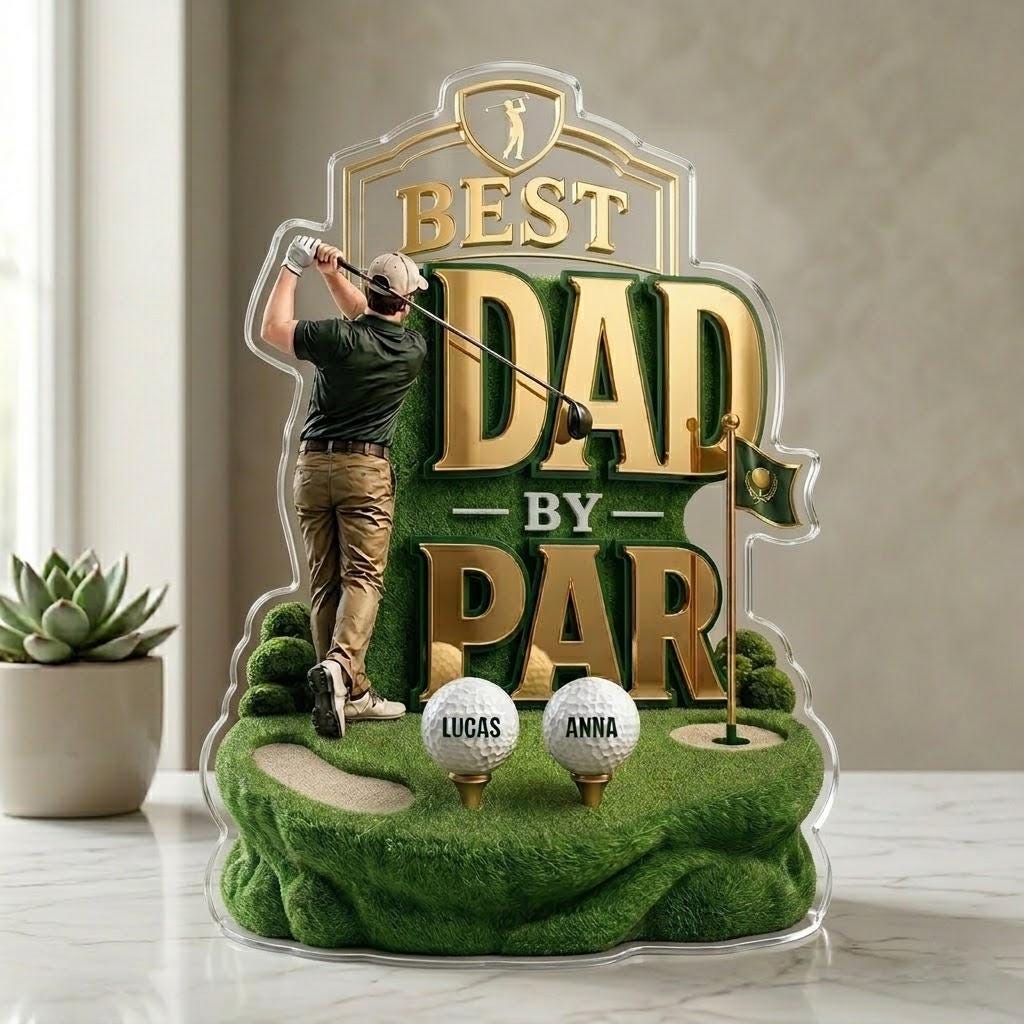 May include: A clear acrylic golf-themed decoration with the words "BEST DAD BY PAR" in gold. It features a golfer figurine, two golf balls with names, and a green golf course design. A small succulent plant is in the background.