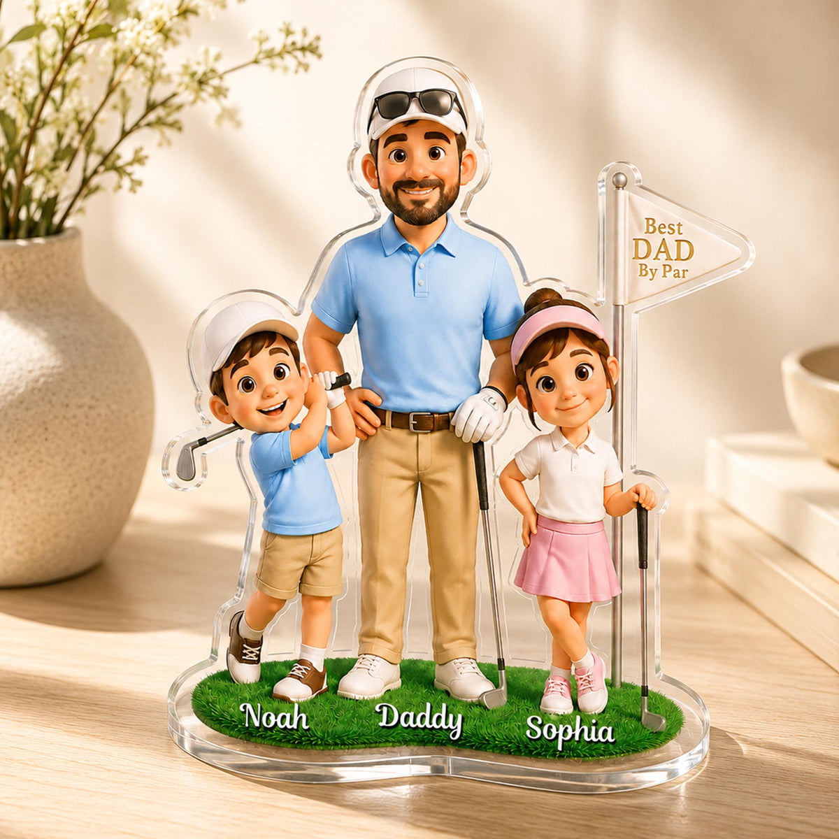 May include: A clear acrylic golf-themed family figurine featuring a father, son, and daughter. The father wears a blue polo shirt and khaki pants. The son and daughter are in golf attire. The base is green with the names Noah, Daddy, and Sophia.