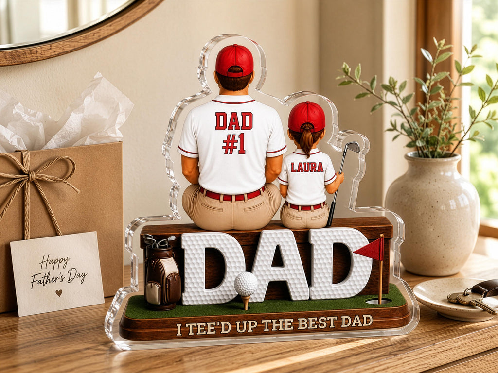 May include: A decorative acrylic display featuring a figurine of a person in a white shirt labeled DAD #1 and a smaller person in a white shirt labeled LAURA, both wearing red caps. The base includes the text DAD and I TEE'D UP THE BEST DAD.