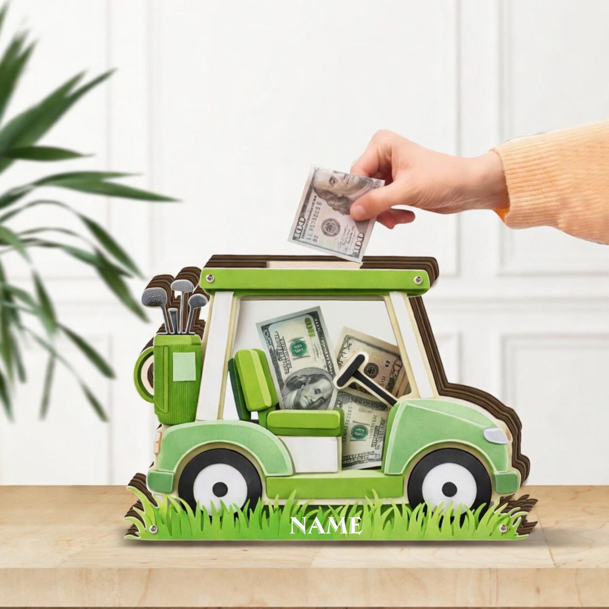 May include: A green and white golf cart-shaped money bank with a brown roof and wheels. The bank is filled with cash, and a hand is inserting a bill. The bank has a golf club holder and a patch of grass with the word "NAME" on it.