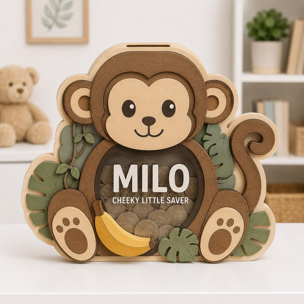 May include: A wooden monkey-shaped piggy bank with a clear front panel displaying coins inside. The design features light and dark brown wood tones, green leaf accents, and a yellow banana. The text MILO and CHEEKY LITTLE SAVER is printed on the front.