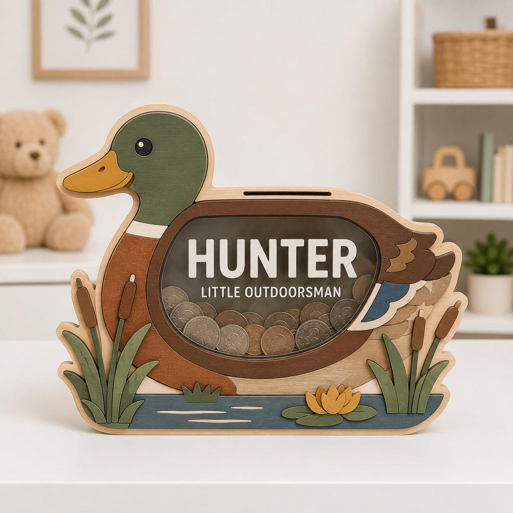 May include: A wooden duck-shaped piggy bank with a clear acrylic window displaying coins inside. The text HUNTER LITTLE OUTDOORSMAN is printed on the window. The duck features green, brown, and yellow tones with cattails and a water lily.