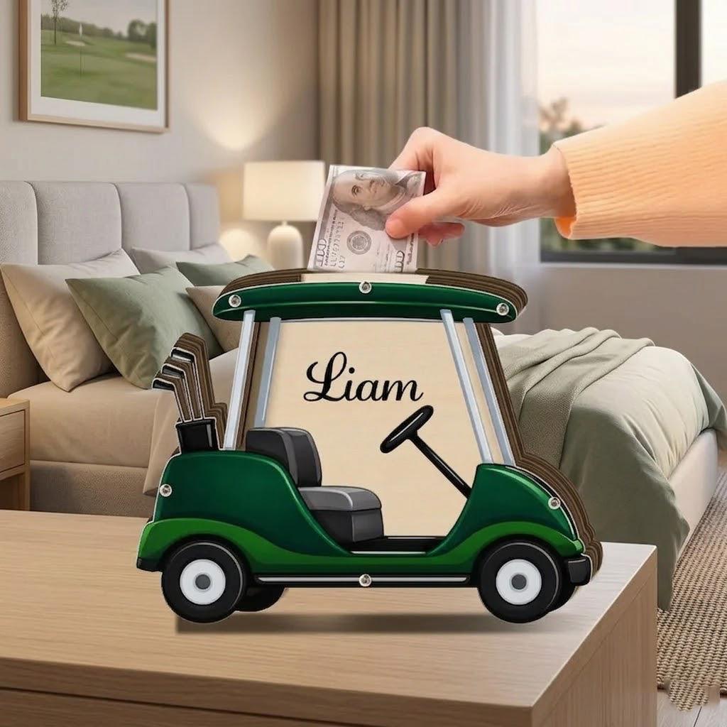 May include: A wooden golf cart-shaped bank in shades of green and brown, with the name "Liam" displayed. The bank has a steering wheel, a seat, and a set of golf clubs. A hand is inserting a U.S. $100 bill into the slot.