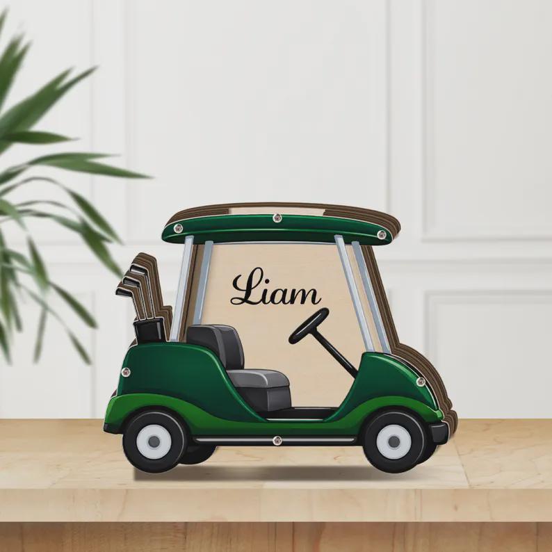 May include: A wooden golf cart-shaped decoration in shades of green, brown, and beige. The cart features a steering wheel, a seat, and a set of golf clubs. The name "Liam" is written on the cart.