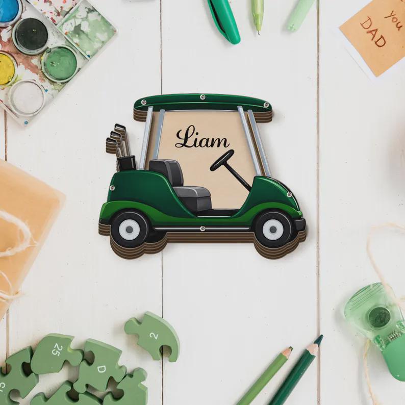 May include: A green golf cart-shaped wooden sign with the name "Liam" in elegant script. The sign includes a steering wheel, golf clubs, and a seat. The cart is set against a neutral background, with other objects like pencils and a puzzle.