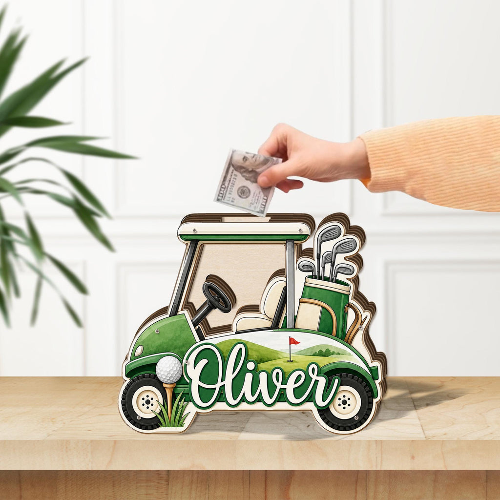 May include: A wooden golf cart shaped money box featuring a green and white color scheme, a golf bag with clubs, a golf ball on a tee, and the name Oliver printed in white script. A slot at the top allows for the insertion of paper currency.