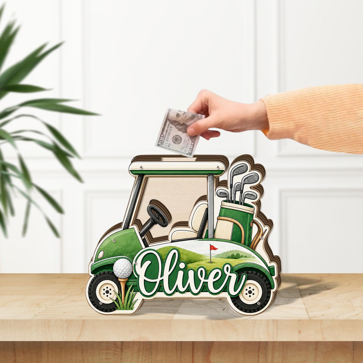 May include: A wooden golf cart shaped money box featuring a green and white color scheme, a golf bag with clubs, a golf ball on a tee, and the name Oliver printed in white script. A slot at the top allows for the insertion of paper currency.