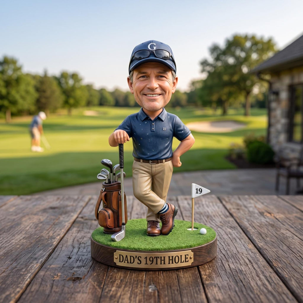 May include: A bobblehead figurine of a golfer wearing a navy blue polo shirt, khaki pants, and a navy cap with a white G. The figure stands on a green base with a golf bag, a club, a flag labeled 19, and a plaque reading DAD'S 19TH HOLE.