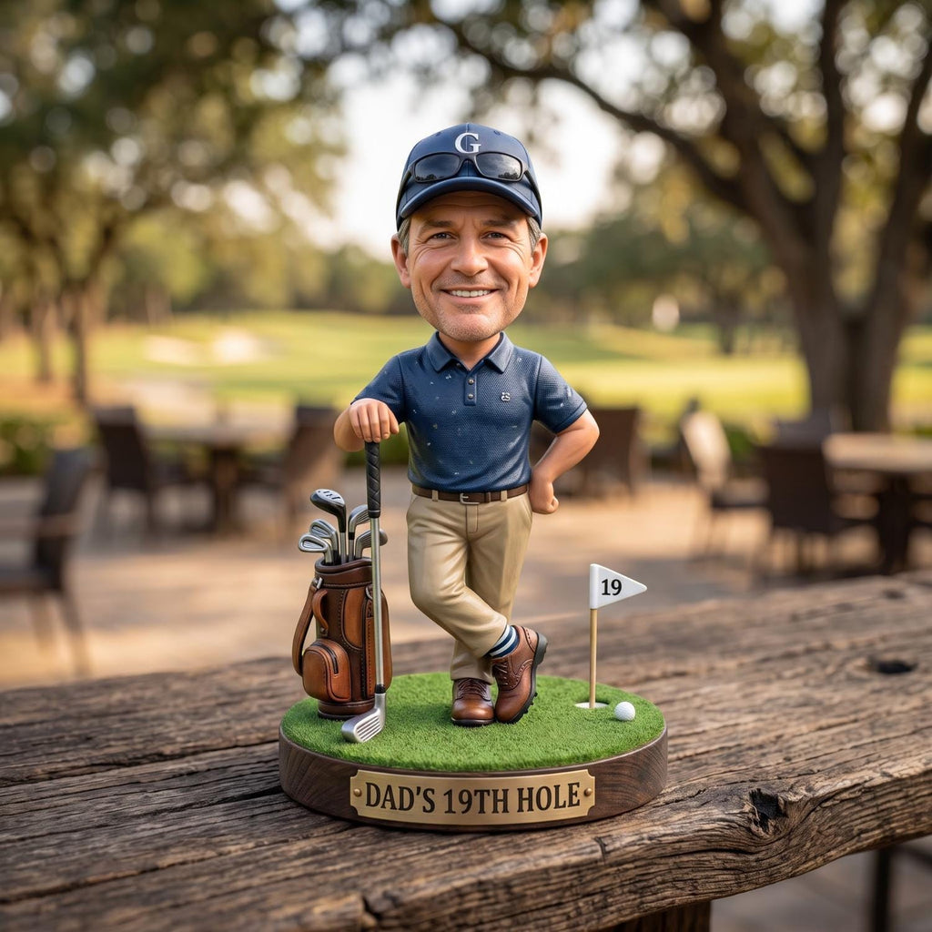 May include: A bobblehead figurine of a golfer wearing a navy blue polo shirt, khaki pants, and a navy cap with a G logo. The figure stands on a green base with a golf bag, a club, a flag marked 19, and a ball. A plaque reads DAD'S 19TH HOLE.