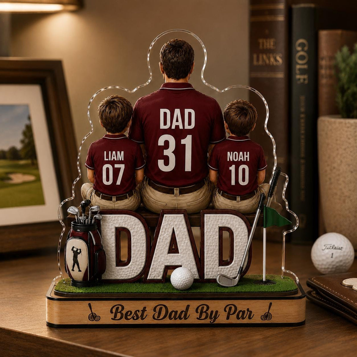 May include: A decorative golf-themed acrylic figurine featuring a father and two sons, with the word "DAD" prominently displayed. The sons' shirts have the names "Liam" and "Noah" with numbers. A golf ball and clubs are also included. The base reads "Best Dad By Par."