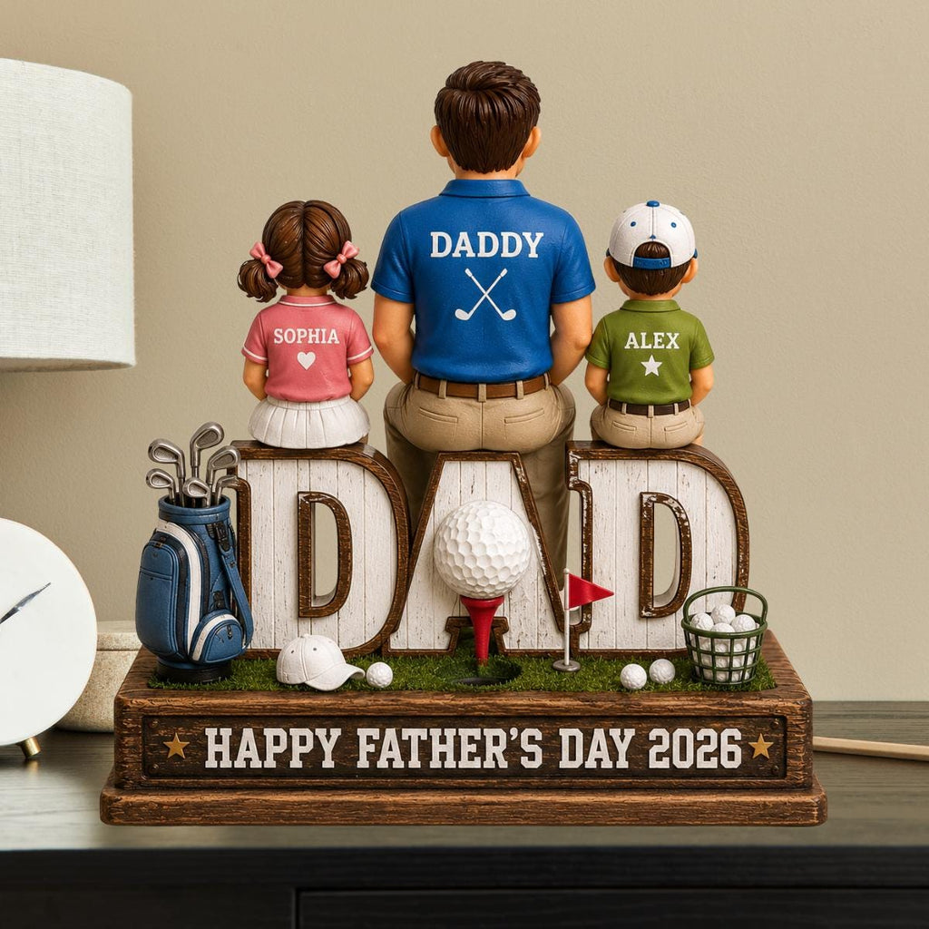 May include: A decorative Father's Day figurine featuring a father and two children, with the word "DAD" in large letters. The father's shirt says "DADDY" and the children's shirts have names. A golf theme is present with a golf ball, clubs, and a basket of balls. The base reads "HAPPY FATHER'S DAY 2026".