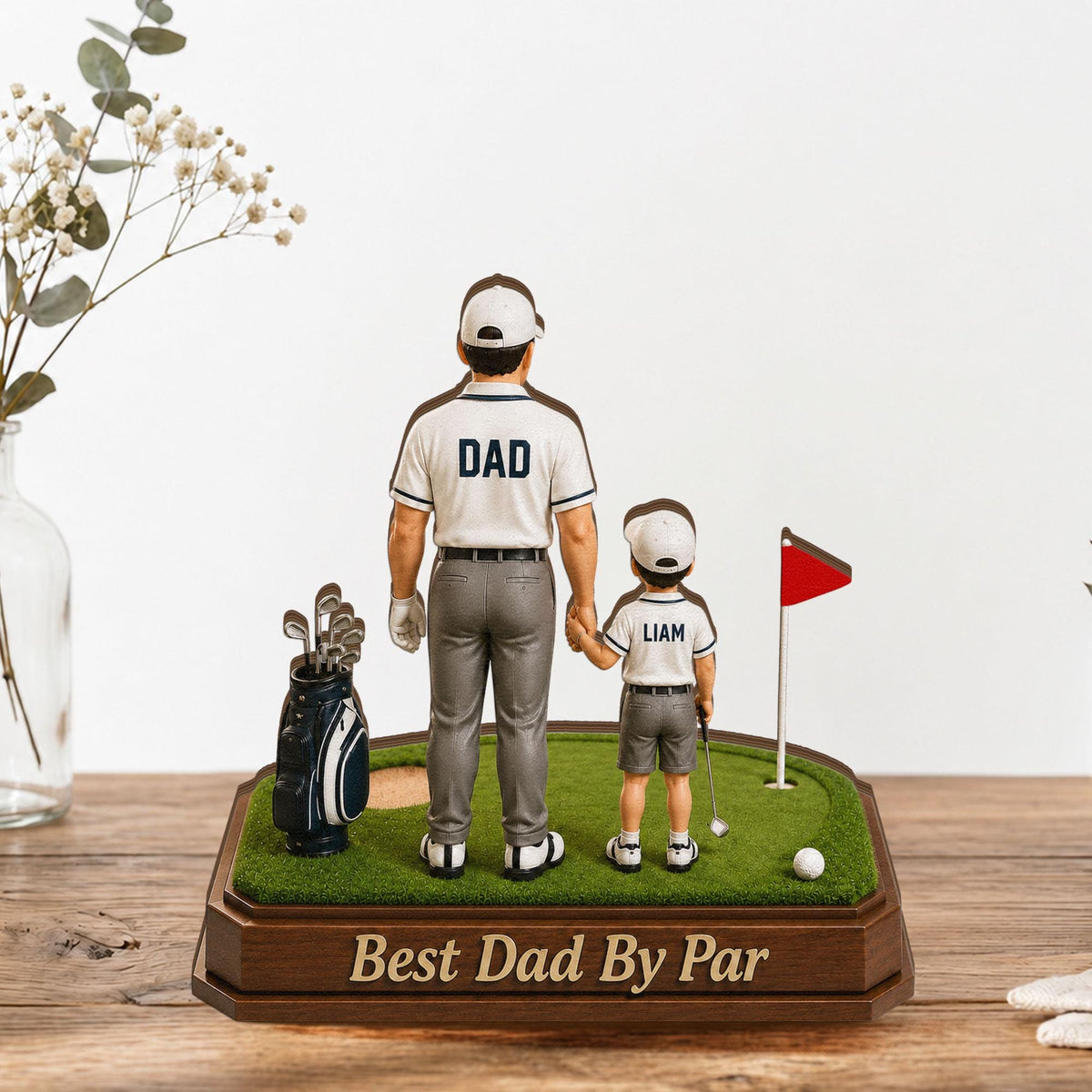 May include: A golf-themed figurine featuring a father and child holding hands on a miniature green. The father's shirt reads "DAD," and the child's reads "LIAM." A golf bag, flag, and the text "Best Dad By Par" are also present.
