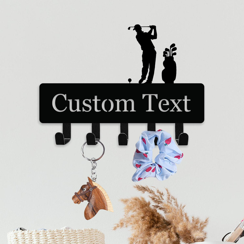 May include: Black metal key holder with hooks, featuring a golfer silhouette and the words "Custom Text". A wooden horse keychain and a blue scrunchie with red strawberries hang from the hooks.