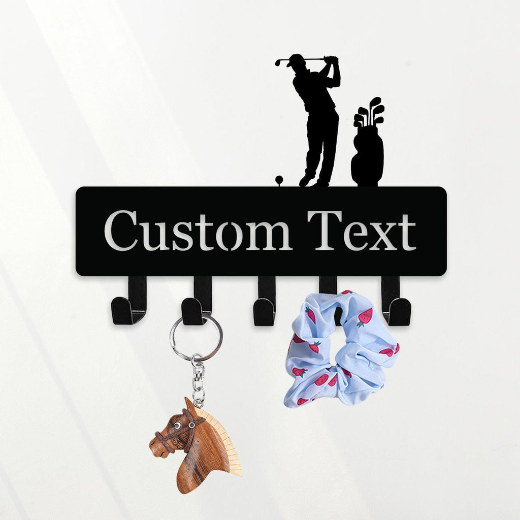 May include: Black metal key holder with a golf theme. The key holder has hooks for keys and accessories. The top features a golfer silhouette and the words "Custom Text". A wooden horse keychain and a blue scrunchie with a strawberry pattern hang from the hooks.