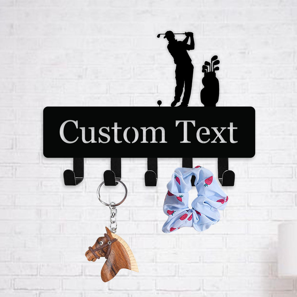 May include: Black metal key holder with a golf theme, featuring a golfer silhouette and the text "Custom Text". Includes five hooks, a wooden horse keychain, and a blue scrunchie with a strawberry pattern.