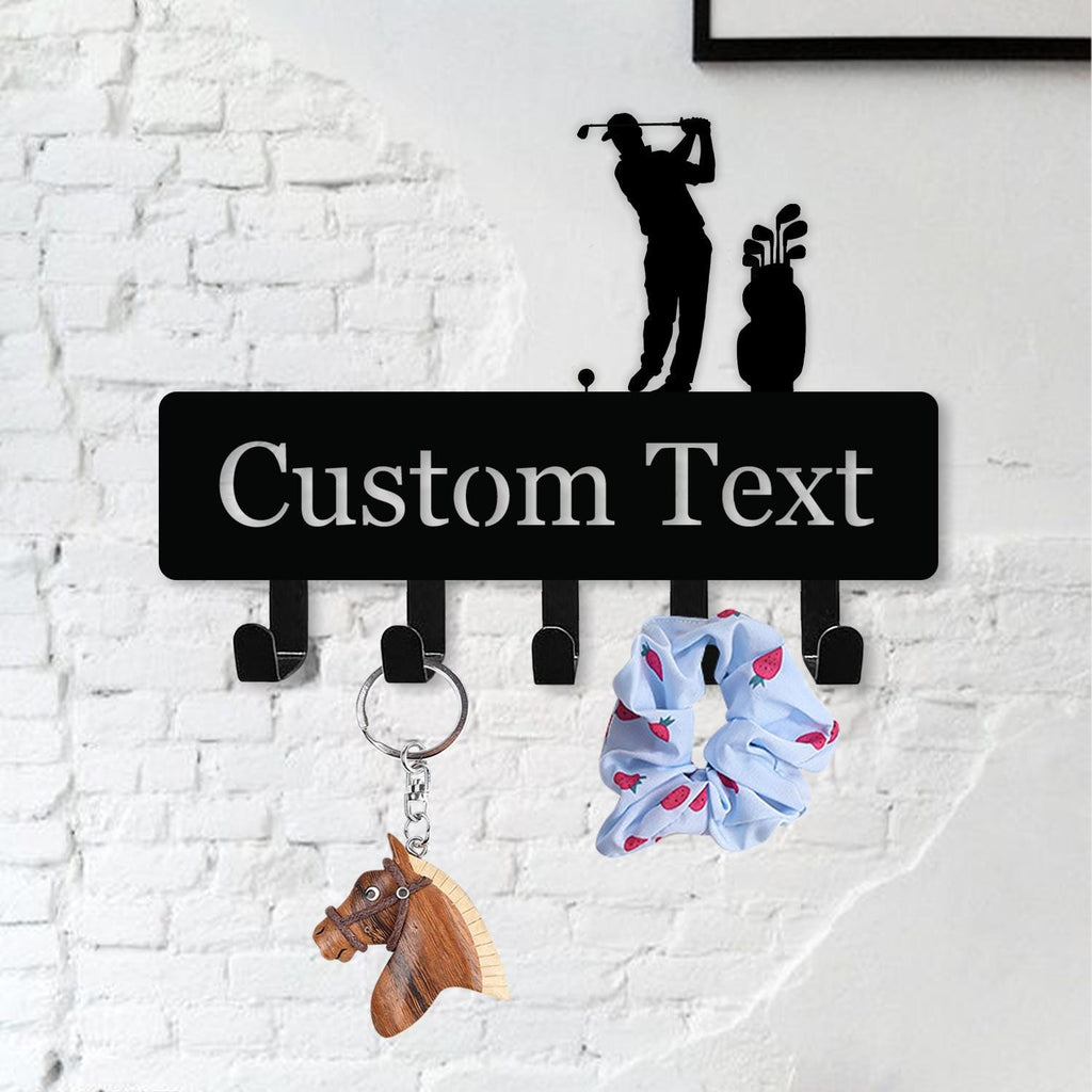 May include: Black metal wall-mounted key holder with the text "Custom Text" in white. The key holder features a golf-themed silhouette and five hooks. A horse keychain and a blue scrunchie with a strawberry pattern hang from the hooks.