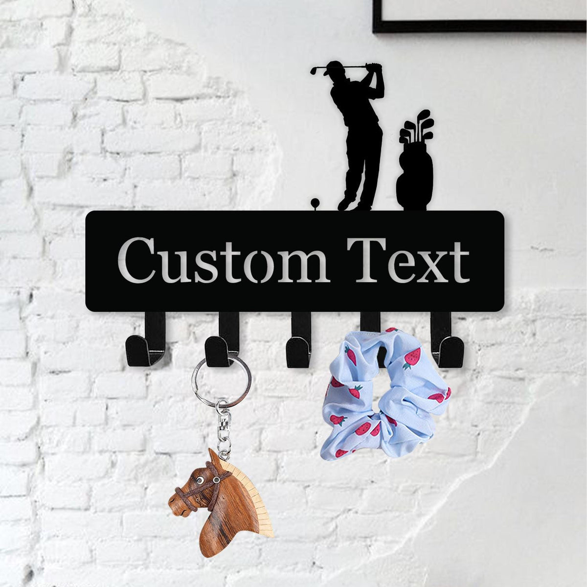 May include: Black metal wall-mounted key holder with the text "Custom Text" in white. The key holder features a golf-themed silhouette and five hooks. A horse keychain and a blue scrunchie with a strawberry pattern hang from the hooks.