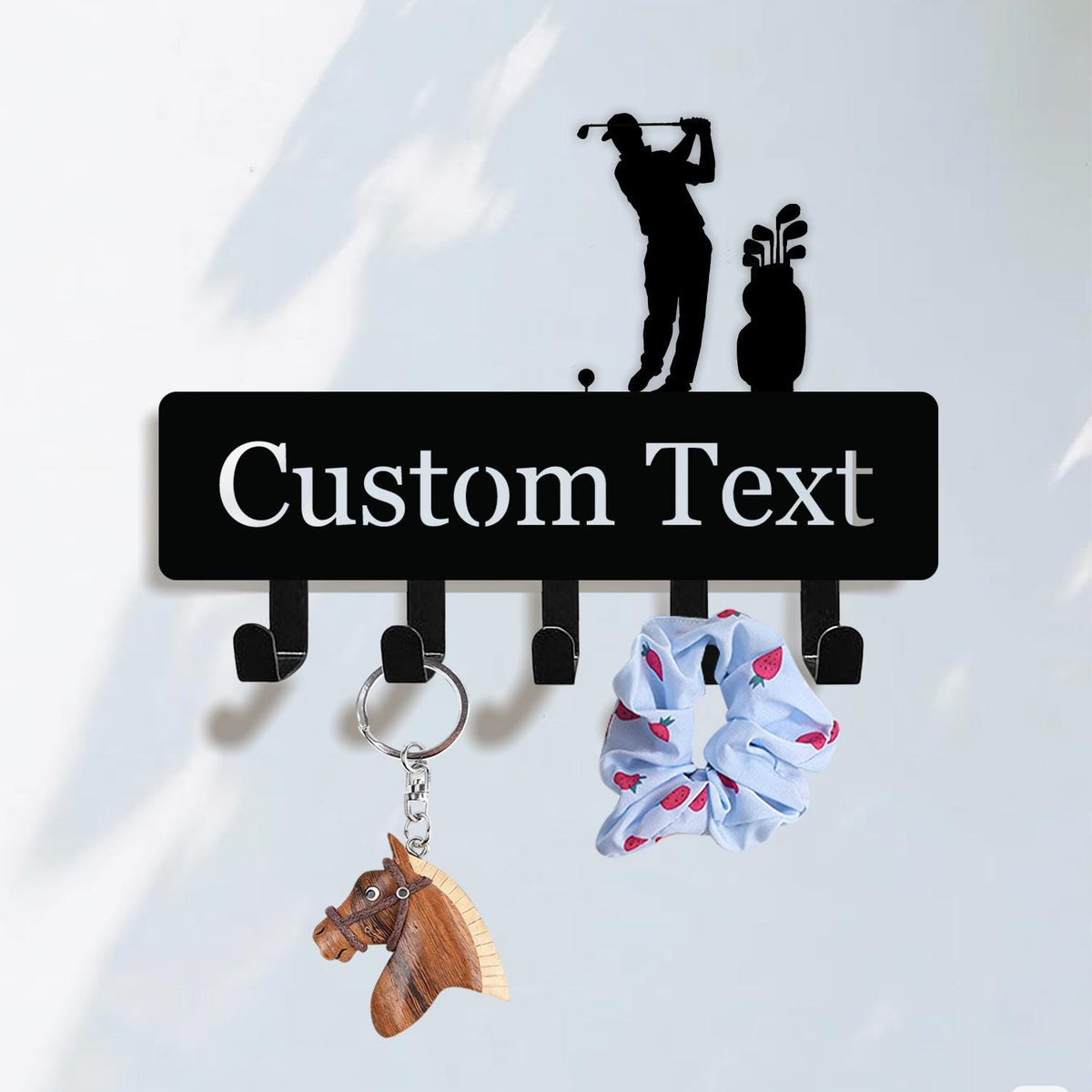 May include: A black metal key holder with five hooks, featuring a golfer silhouette and the words "Custom Text" in white. A wooden horse keychain and a blue scrunchie with red strawberries hang from the hooks.