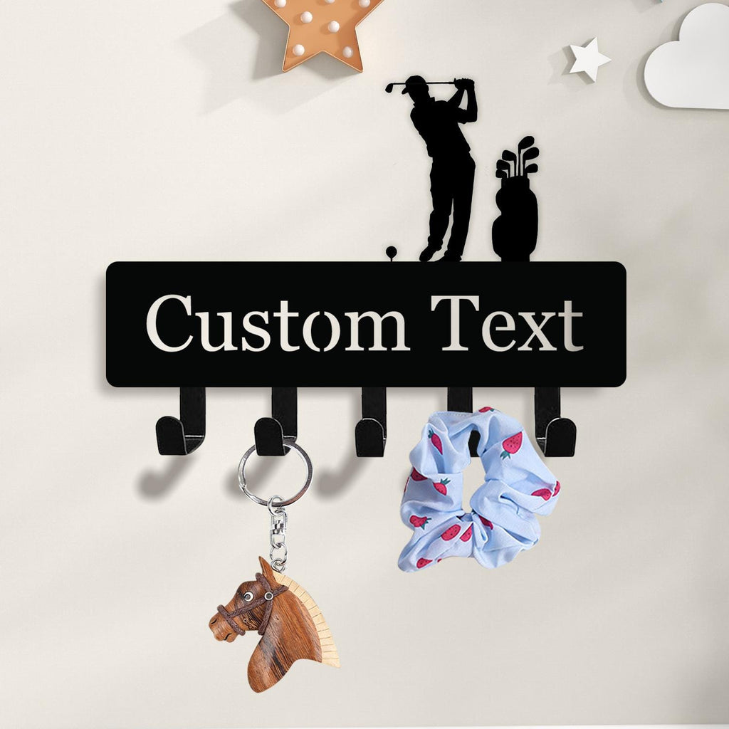 May include: Black metal wall-mounted key holder with hooks, featuring a golf-themed silhouette and the text "Custom Text". A wooden horse keychain and a blue scrunchie with a strawberry pattern hang from the hooks. Decorative stars and clouds adorn the wall.