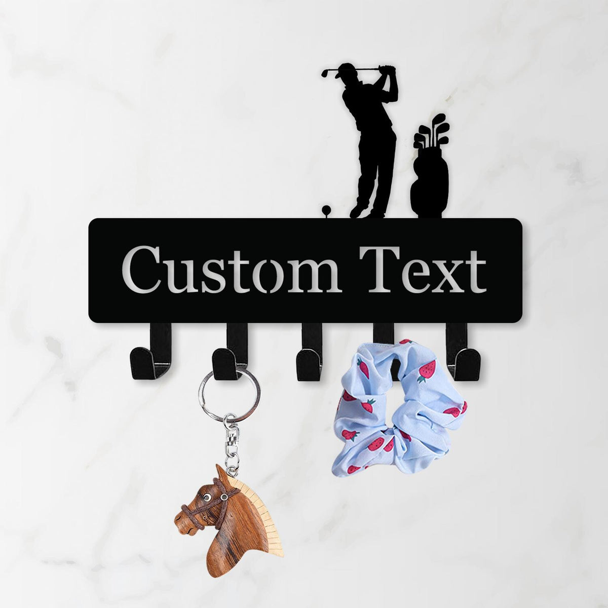 May include: Black metal wall-mounted key holder with a golf theme, featuring a golfer silhouette and golf bag. The holder has hooks for keys and accessories, with the text "Custom Text". A wooden horse keychain and a blue scrunchie with red strawberries are hanging from the hooks.