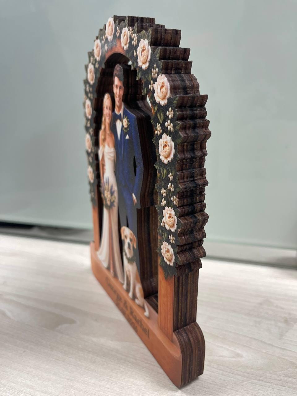 May include: A multi-layered wooden wedding ornament featuring a cutout of a couple and a dog standing under a floral arch. The arch is decorated with painted white roses and green leaves. The base is engraved with the names George and Rachel.