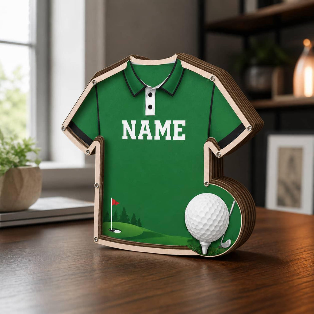 May include: A green wooden shadow box shaped like a polo shirt with the word NAME printed in white on the chest. The design features a golf course graphic with a red flag and a white golf ball resting on a tee in the lower right corner.