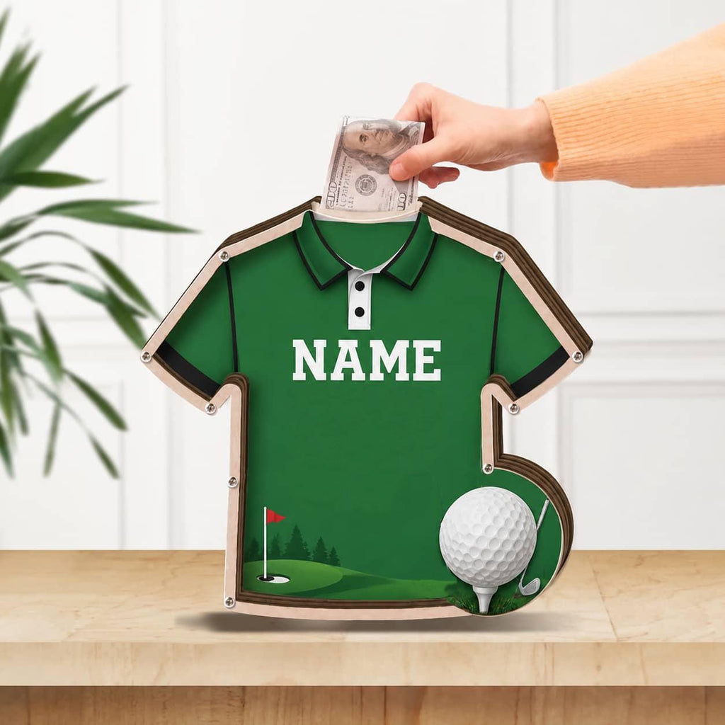 May include: A green wooden money box shaped like a polo shirt with a golf theme. The front features the word NAME in white text, a golf course graphic with a flag, and a 3D golf ball. A slot at the top allows for the insertion of paper currency.