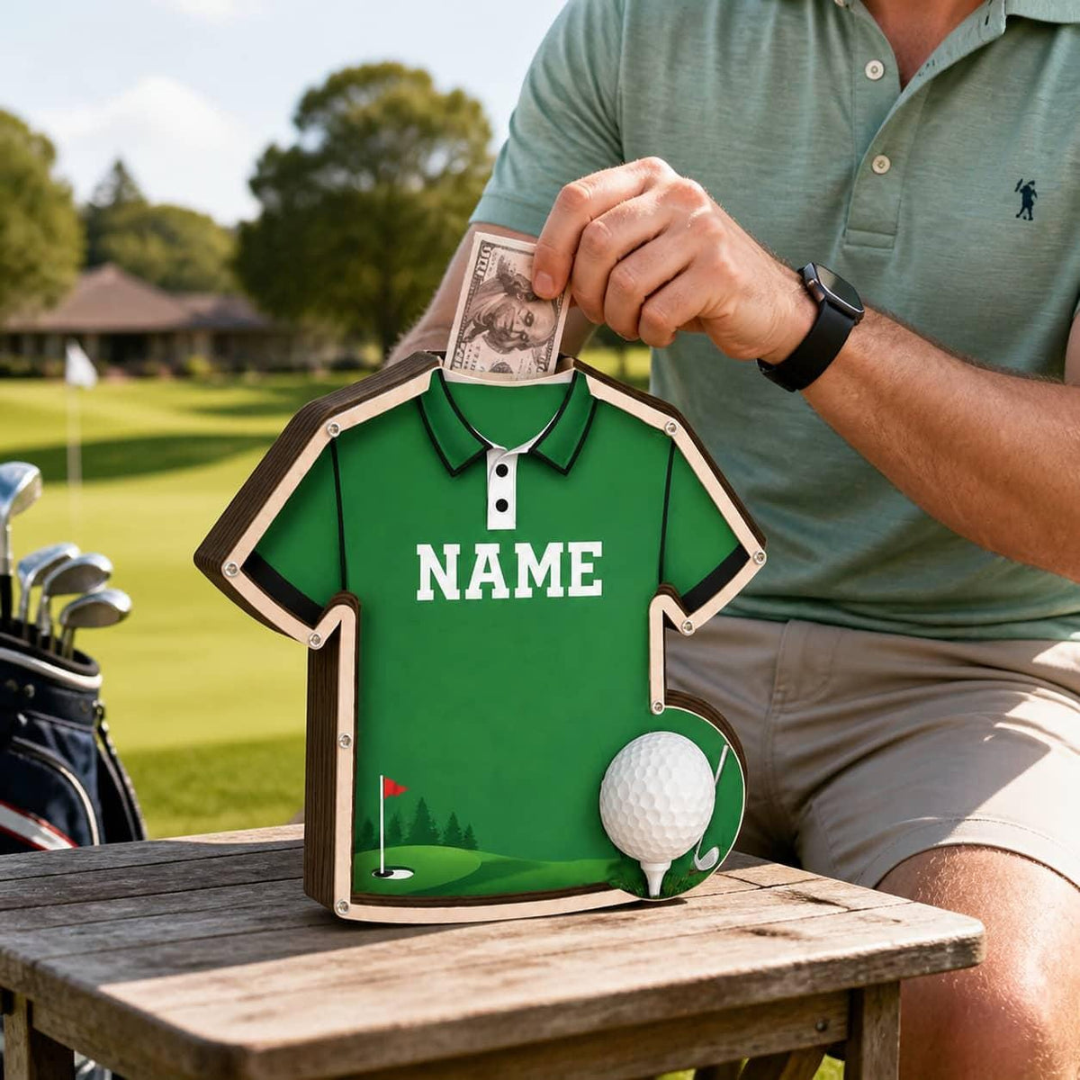 May include: A green wooden money box shaped like a polo shirt with the word NAME printed in white across the chest. The design features a golf course graphic at the bottom, a white golf ball accent, and a slot at the collar for inserting paper money.