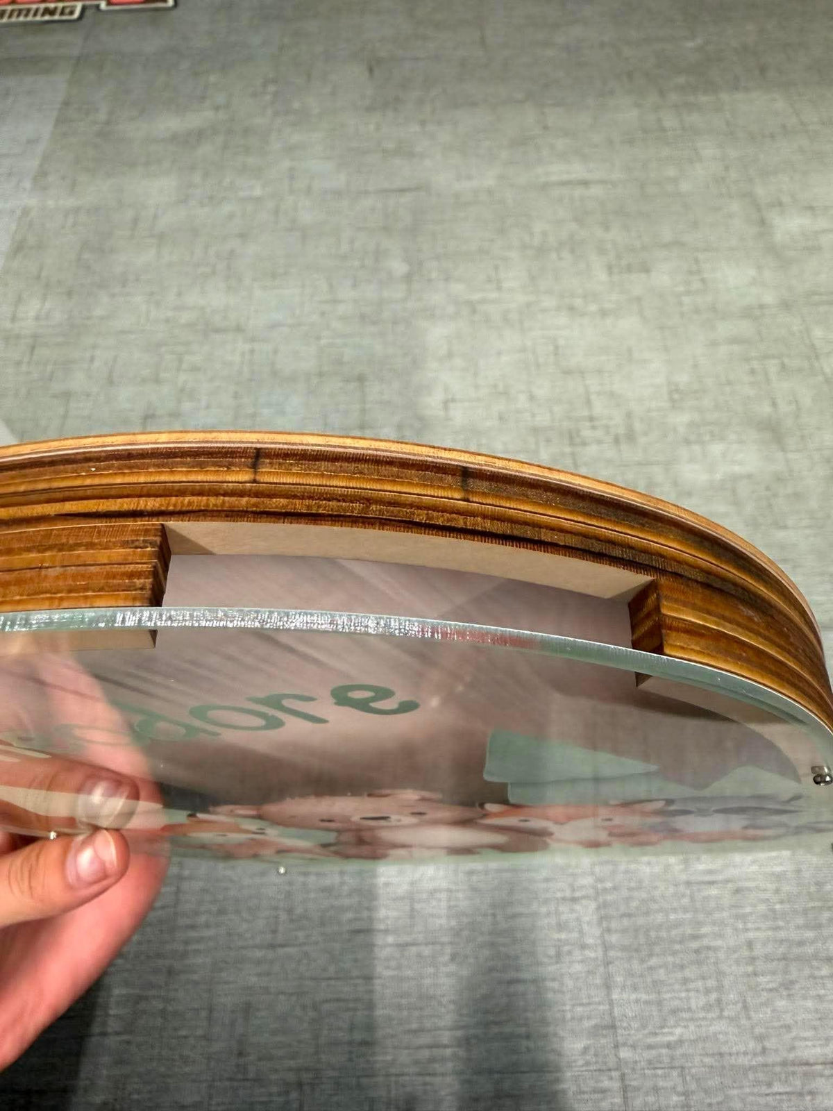 May include: A circular wooden frame with a layered, stacked edge design. A clear acrylic panel is inset into the wood, featuring a printed graphic of small woodland animals and the partial text "adore" in a light green, sans-serif font.