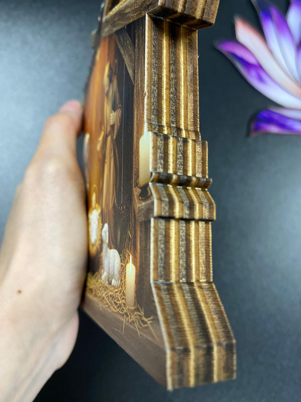 May include: A layered wooden plaque featuring a printed nativity scene with a central figure, a lamb, and a lit candle. The side profile reveals the stacked, dark-edged wood construction with a stepped, architectural design and a smooth, flat front surface.
