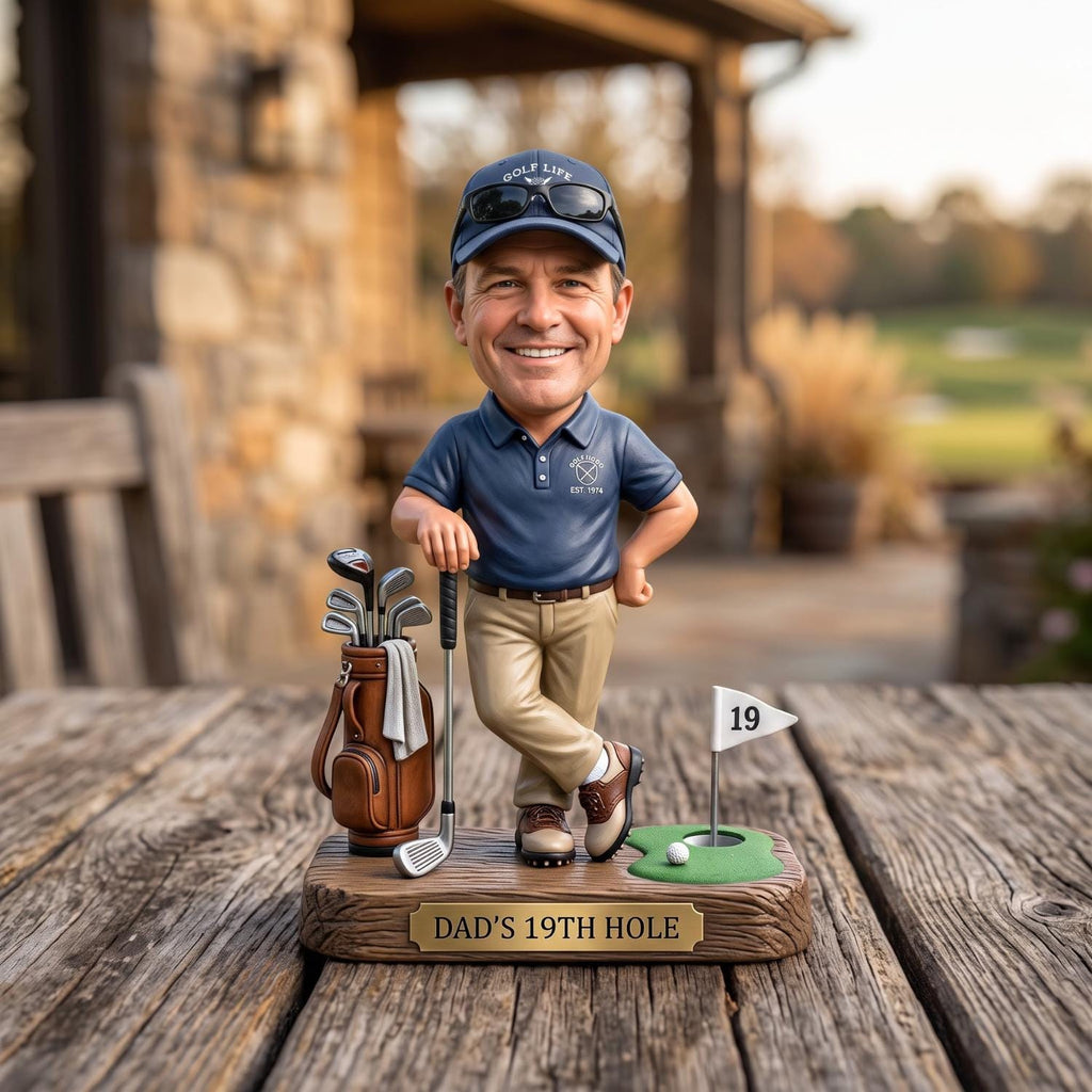 May include: A bobblehead figurine of a golfer wearing a navy blue polo shirt, khaki pants, and a navy cap with sunglasses. The figure stands on a base labeled DAD'S 19TH HOLE, featuring a golf bag with clubs, a putter, and a green with a flag marked 19.
