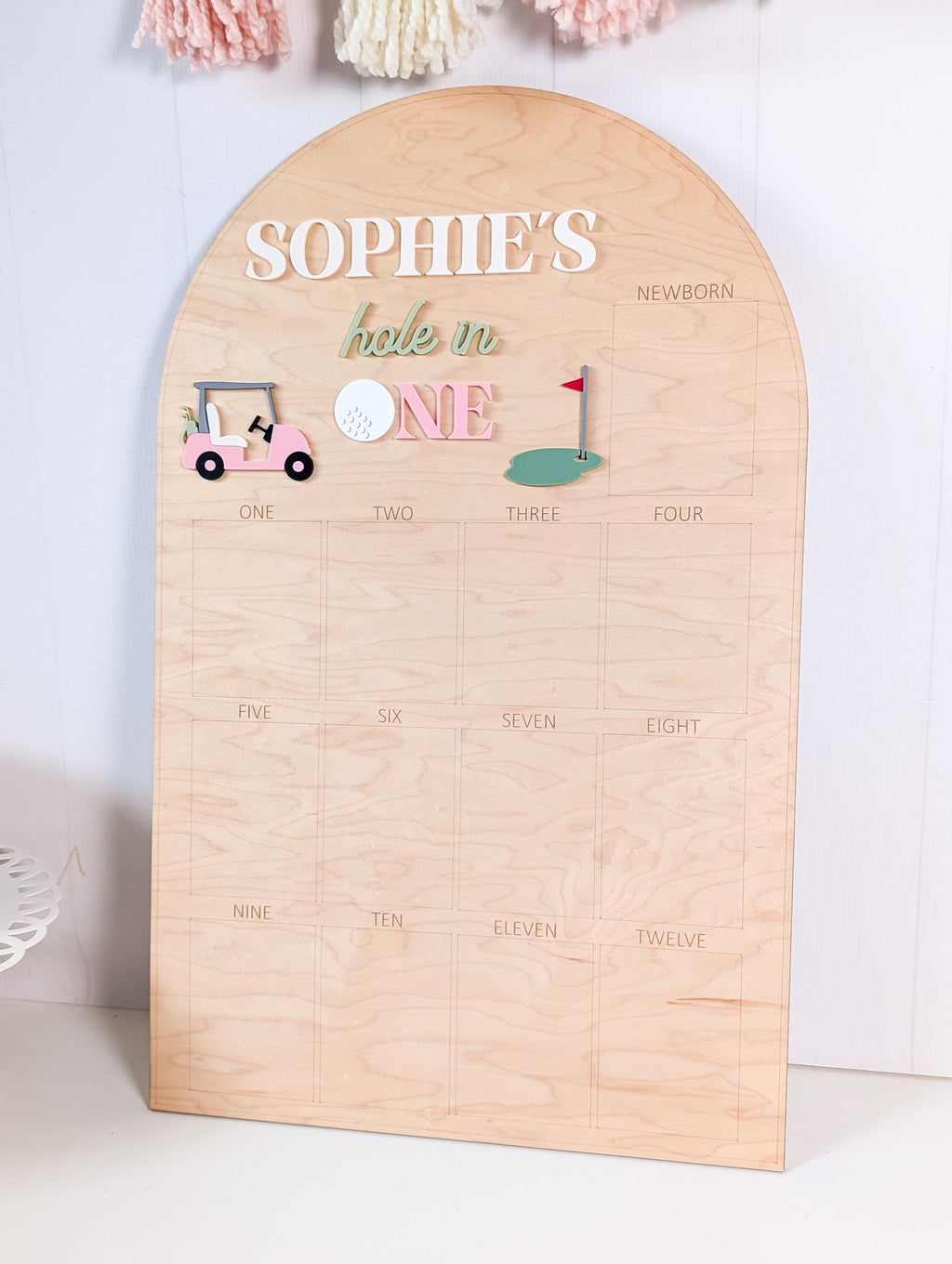 May include: A wooden milestone board with an arched top. The board features the name "Sophie's" and the phrase "hole in one" in decorative lettering. Golf-themed embellishments include a pink golf cart, a golf ball, and a green with a flag. The board is divided into sections for tracking milestones.