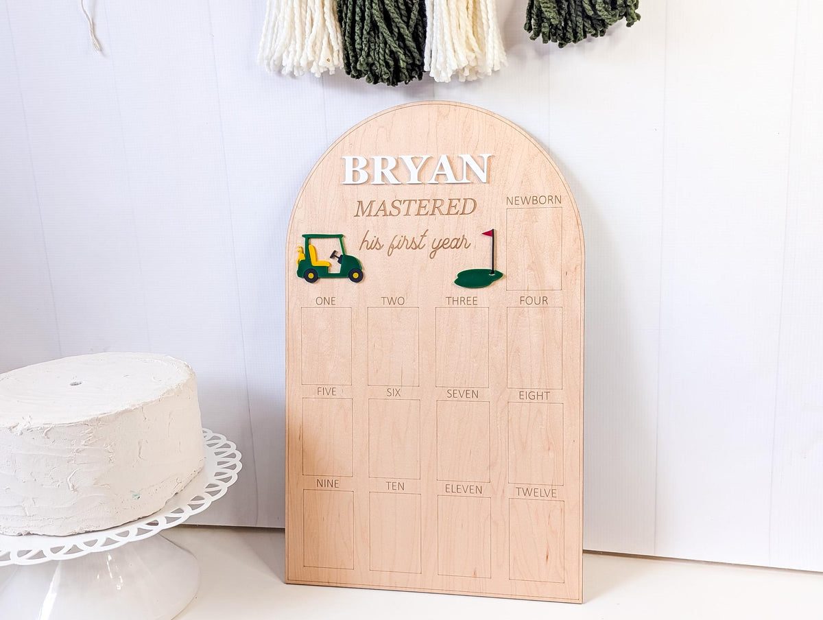 May include: A wooden milestone board with the name "BRYAN" at the top. The board has a golf theme with a golf cart and a flag. It includes the text "MASTERED his first year" and numbered boxes for each month.