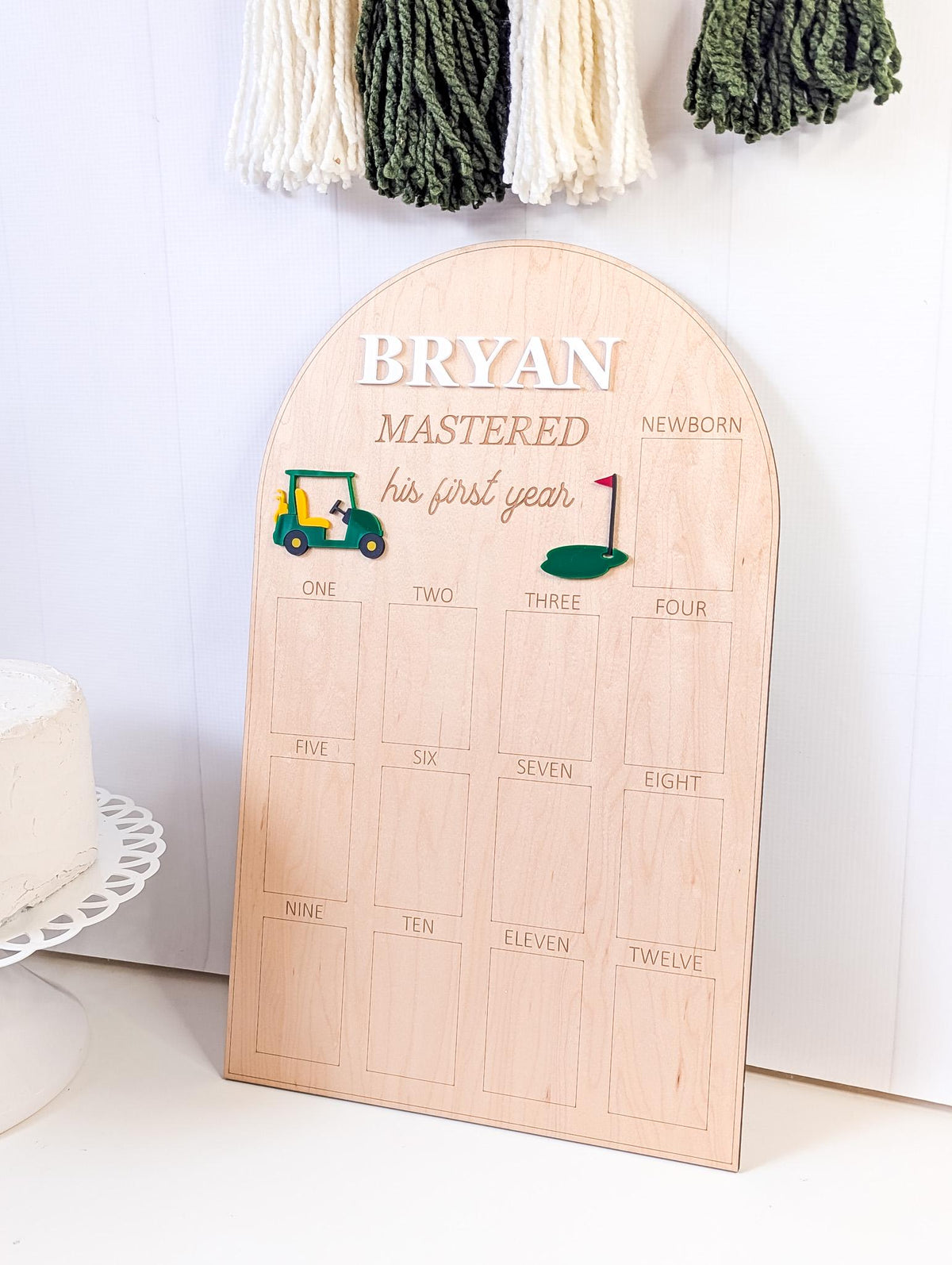 May include: A wooden milestone board with the name "BRYAN" in white letters. The board has a golf theme with a golf cart and flag graphic. It includes spaces for months one through twelve, and the text "MASTERED his first year".