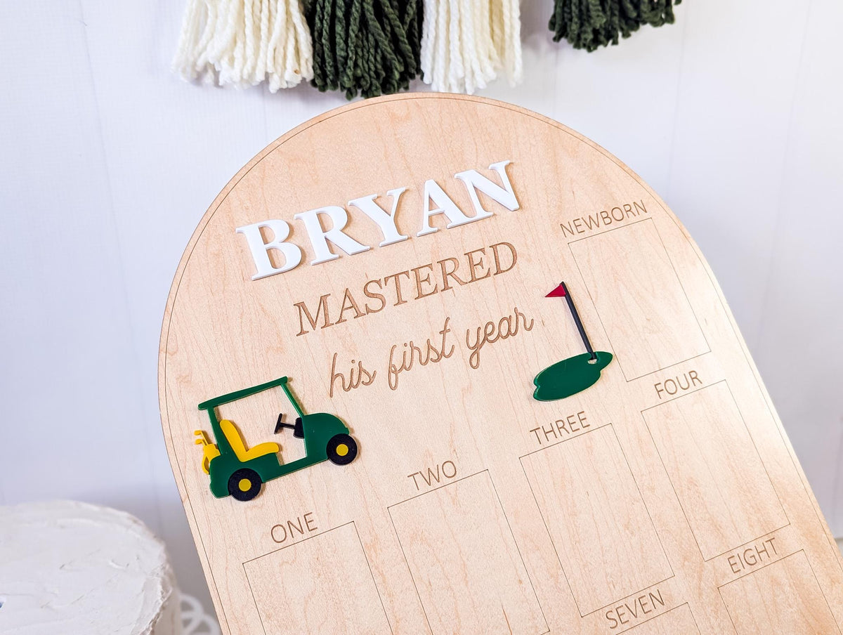May include: Wooden milestone board with the name "BRYAN" in white letters. The board has a golf theme, with a green and yellow golf cart and a golf flag. The text "MASTERED his first year" is engraved in a script font.