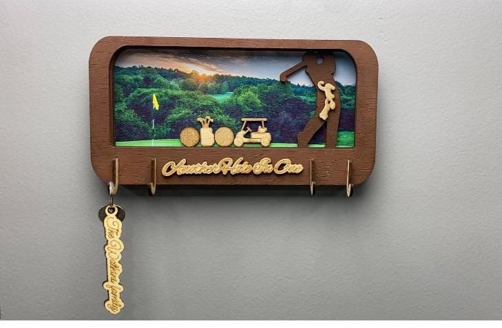 May include: Wooden key holder with a golf theme. The frame is brown and features a golf course scene with a golfer silhouette. The text "Another Hole in One" is displayed, along with a key with the text "The [Last Name] Family".