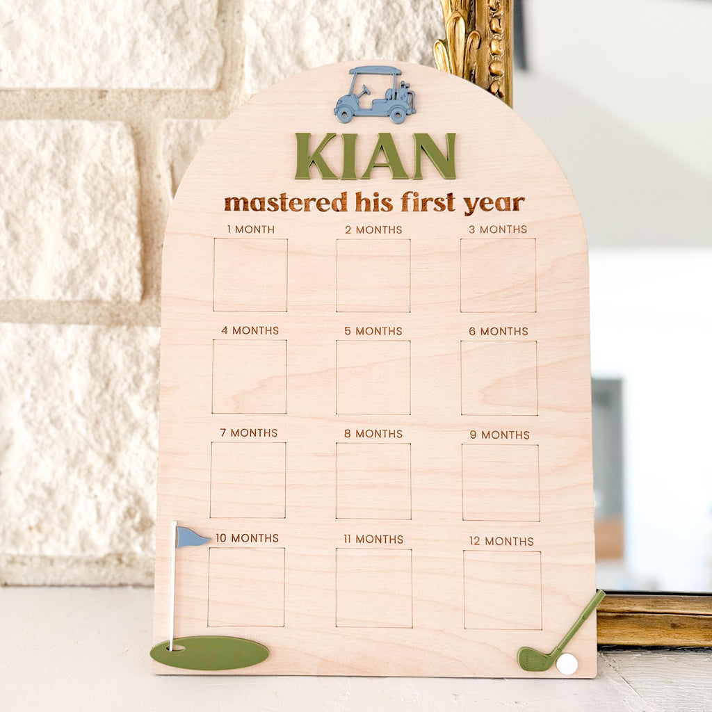 Personalized wood golf first birthday milestone photo board with 12 spaces for monthly 2x2 baby photos, featuring custom name in acrylic and golf accents, displayed upright