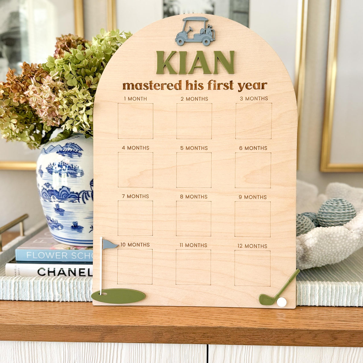 personalized golf-themed milestone board with baby’s monthly photo and name in acrylic, styled as first party decor on a table with blue accents