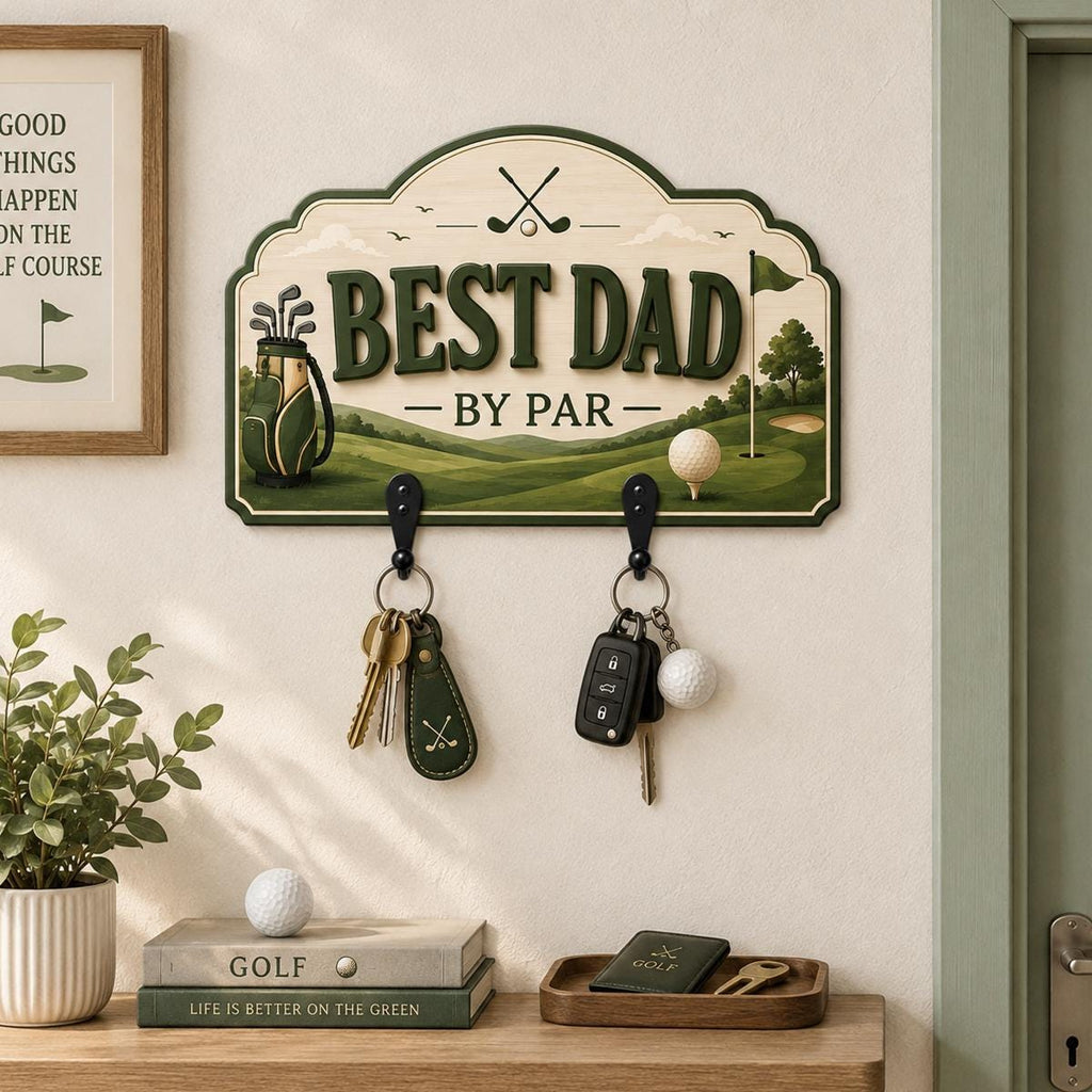 May include: A wall-mounted wooden key holder featuring a golf course illustration, the text BEST DAD BY PAR, and two black metal hooks. The design includes a golf bag, a flag, and a golf ball. Two sets of keys hang from the hooks below the sign.