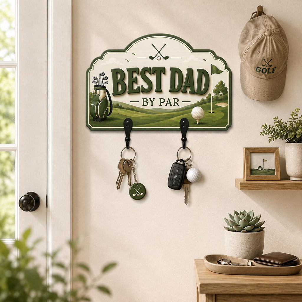 May include: A wall-mounted wooden key holder with a golf theme, featuring the text BEST DAD BY PAR. The design includes a golf bag, a golf ball, and a flag on a green course. Two black metal hooks hold sets of keys, including a golf-themed keychain.