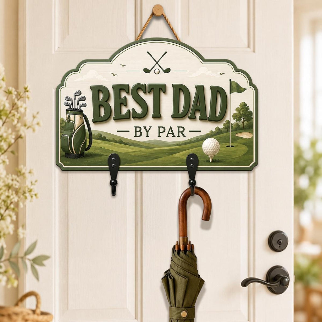 May include: A decorative wall sign with a golf theme, featuring the text BEST DAD BY PAR, a golf bag, and a golf ball on a tee. The sign has a cream background with green accents and includes two black metal hooks for hanging items like an umbrella.